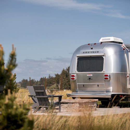 A shiny silver trailer parked outdoors with chairs and a fire pit nearby under a blue sky, surrounded by greenery.