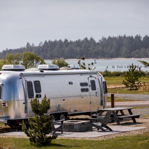 A silver Airstream trailer is parked near a picnic table by a scenic lakeside, with trees and a distant forest in the background.