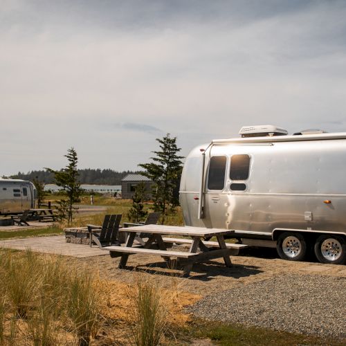 The image shows two silver trailers parked in a campground with picnic tables and grassy surroundings under a partly cloudy sky.