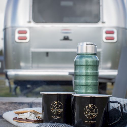 Two black mugs, a green thermos, and snacks on a plate, set on a blanket, with a shiny trailer in the background.