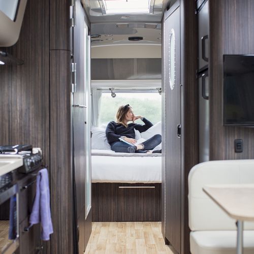 A person is relaxing on a bed inside a modern RV, featuring a kitchenette and seating area with sleek, dark wood cabinetry.