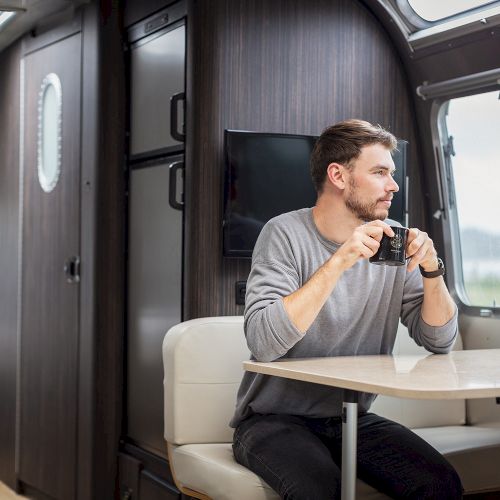 A person sits inside a modern RV, holding a mug and looking out the window. The interior features sleek, dark wood paneling and a cozy atmosphere.