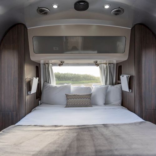 This image shows a cozy bed inside an Airstream trailer with wood paneling, soft pillows, and windows on both sides.