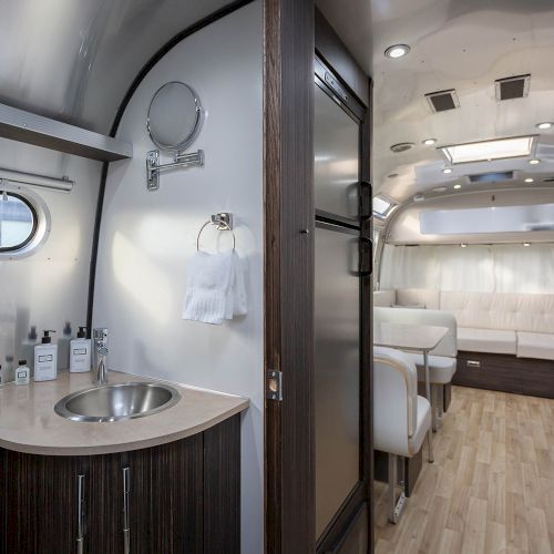 Interior of a modern camper with a sink, seating area, kitchenette, and wood flooring, featuring sleek finishes and ample natural light.