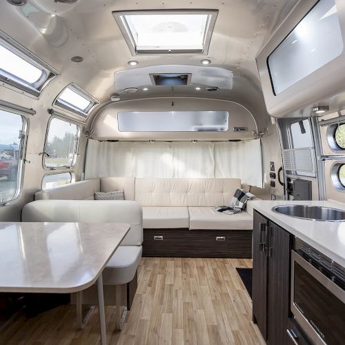 This image shows the interior of a modern Airstream trailer with a kitchen, table, couch, wooden flooring, and large windows.