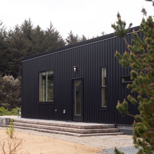 A modern black house with large windows is surrounded by trees and a small patio area.