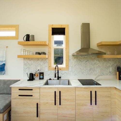 A modern kitchen with wooden cabinets, open shelves, a stove, sink, kettle, and a hood. Small window above the sink. Clean design.