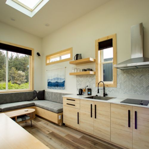 A cozy modern kitchen and living area with light wood cabinets, a gray sofa, and large windows overlooking greenery.