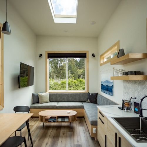 A modern tiny house interior with a skylight, compact kitchen, corner sofa, wall-mounted TV, and a view of trees through large windows.