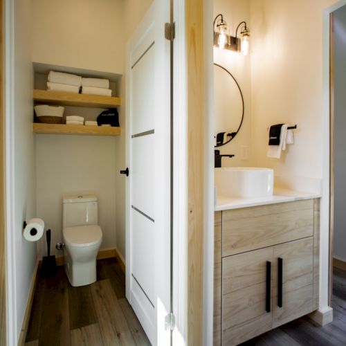 This image shows a modern bathroom with a toilet, open shelves with towels, a vanity with a round mirror, and wall-mounted lights.