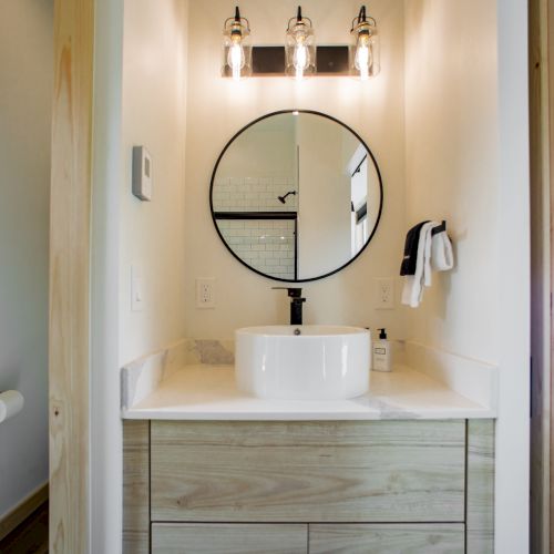 A modern bathroom with a round mirror, white basin, wood cabinet, and three-bulb light fixture. Towel and toiletries are on the sink.