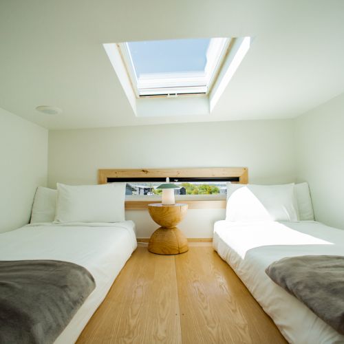 The image shows a bright room with two beds, a small table in between, and a skylight above, allowing natural light to fill the space.