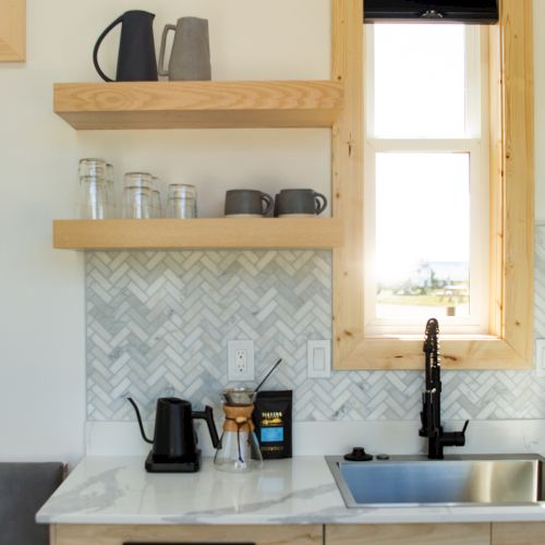 A modern kitchen scene with wooden shelves, a window, kettles, jars, a coffee maker, black faucet, and herringbone backsplash.