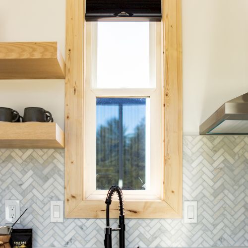 A kitchen sink with a black faucet is beneath a window with wood trim. To the left are shelves with cups and a herringbone tile backsplash.