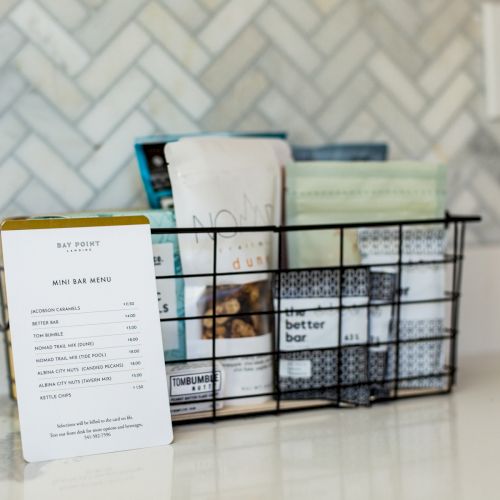 A wire basket on a countertop holds various packaged snacks. A small menu listing snack bar options and prices is propped in front.