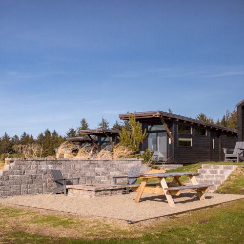 The image shows a scenic setting with wooden cabins, outdoor seating, picnic tables, and neatly landscaped surroundings under a clear blue sky.