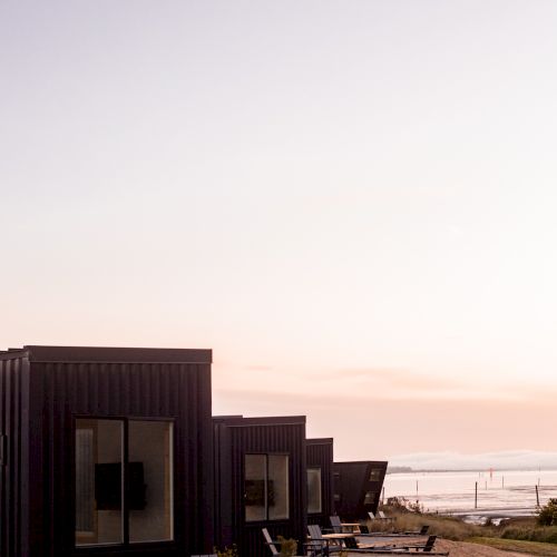 The image shows a row of modern, small, black cabins next to a sandy area with a view of the sea under a clear sky.