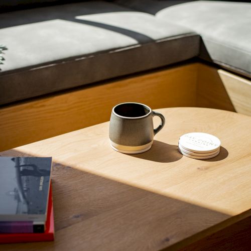 A cozy seating area with a wooden table holding a black mug, a white coaster, and a couple of books, lit by sunlight.