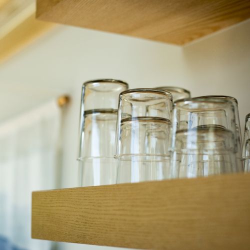 Several clear glass cups are placed upside down on a wooden shelf in a bright room.