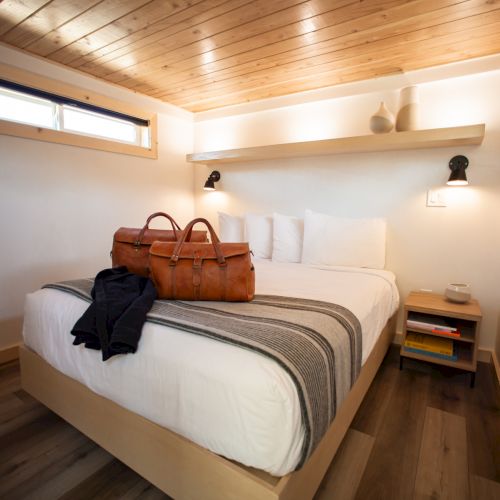 A cozy bedroom with a wooden ceiling, a bed with striped bedding, two bags, a shelf, lamps, and a side table with books and a cup.