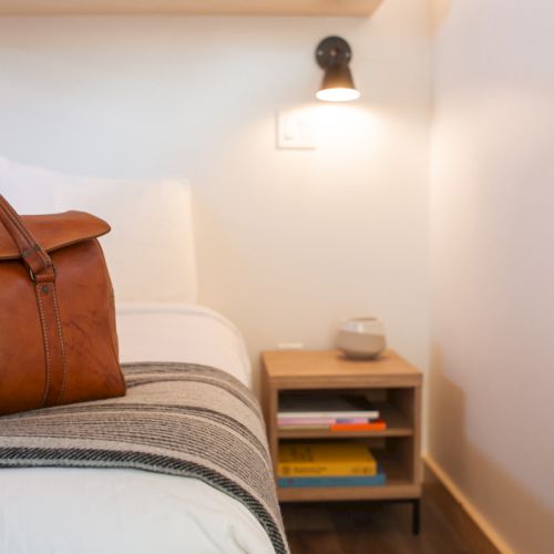 A leather bag on a bed, next to a nightstand with books and a lamp on the wall.