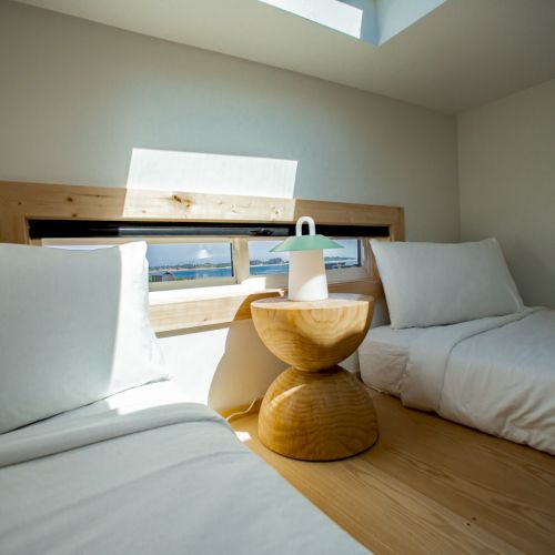 The image shows a minimalist bedroom with two beds, a wooden side table, and soft lighting from a skylight.