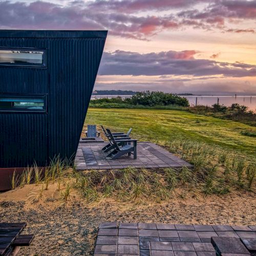 Modern house by the beach with a wooden deck, outdoor seating, and a scenic sunset view.