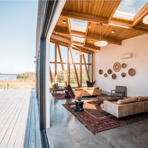 A modern living room with wooden ceilings and skylights opens to a deck, featuring sofas, armchairs, and wall decor on a patterned rug.