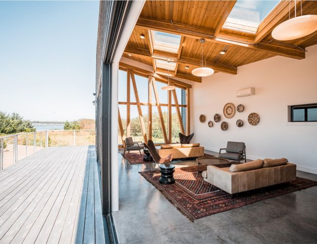 A modern living room with wooden ceilings and skylights opens to a deck, featuring sofas, armchairs, and wall decor on a patterned rug.