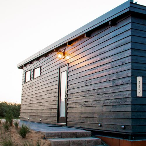 A modern, small house with dark wood siding, a sloped roof, and steps leading to the door. There’s a light fixture above the entrance.