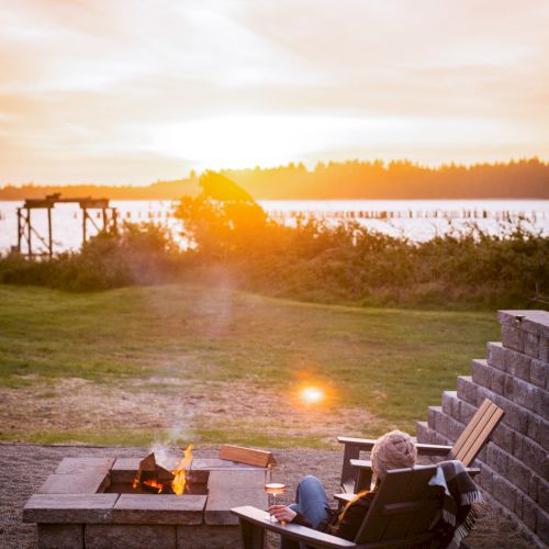 A person sits in an Adirondack chair near a fire pit, overlooking a scenic lake at sunset.