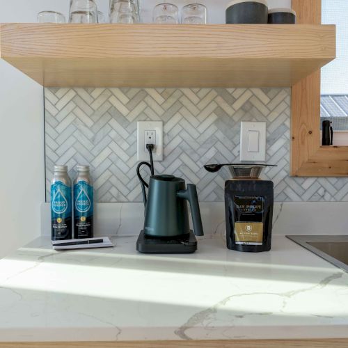 A kitchen counter with a kettle, coffee bags, and thermoses, under a shelf with glasses, near a sink with a herringbone tile backsplash.