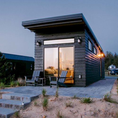 A modern tiny house with large windows, outdoor seating, and steps, set in a natural landscape, with other structures and trailers nearby.