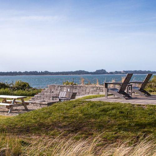 Outdoor seating area with chairs and a picnic table overlooking a lake, surrounded by grass and shrubs under a clear sky.