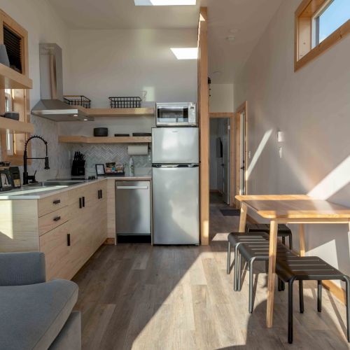 The image shows a modern kitchen with wooden cabinets, a fridge, a stove, a sink, a small dining table with stools, and natural lighting.