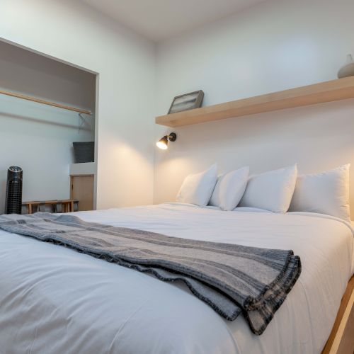 A modern bedroom with a double bed, a striped throw, a floating shelf, wall-mounted lights, and an open closet. Minimalist decor completes the look.