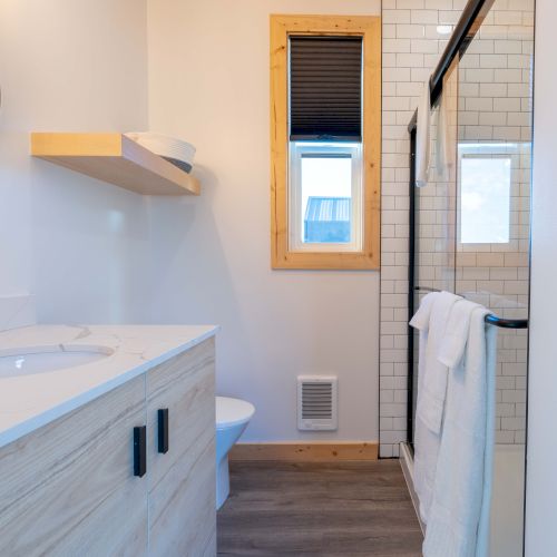 The image shows a modern bathroom with a sink, wooden cabinets, a mirror, a shower, white tiles, and a small window with a blind.