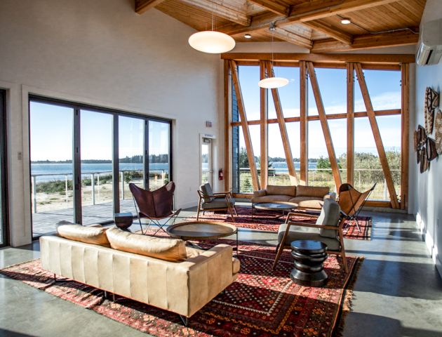 A modern living room with wooden beams, large windows, and stylish furniture on patterned rugs, overlooking an outdoor view.