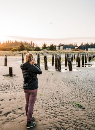 A person stands on a sandy beach taking photos, with wooden posts in the background and a sunset sky.