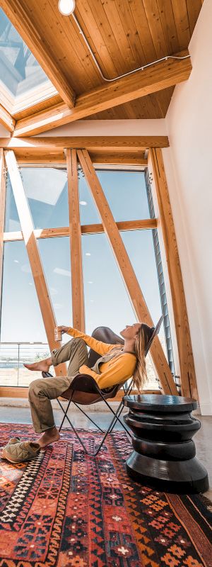 A person relaxes in a modern room with a large wooden window, geometric ceiling, and a patterned rug.