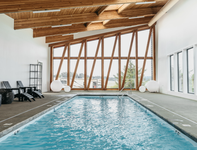 An indoor pool area with a modern wooden ceiling and large triangular windows, featuring lounge chairs and spherical lights.