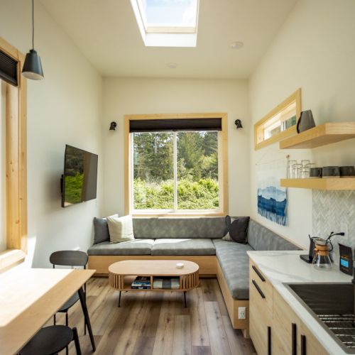 A cozy, modern tiny home interior with skylight, kitchen, dining table, TV, and sofa by a window overlooking greenery.