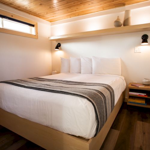 A cozy bedroom with a wooden ceiling, white bed, striped blanket, bedside table, books, a cup, and soft lighting adorn the space.