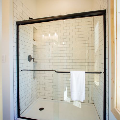 The image shows a modern shower with white subway tiles, a black metal frame, and a glass door, featuring a towel on the handle.