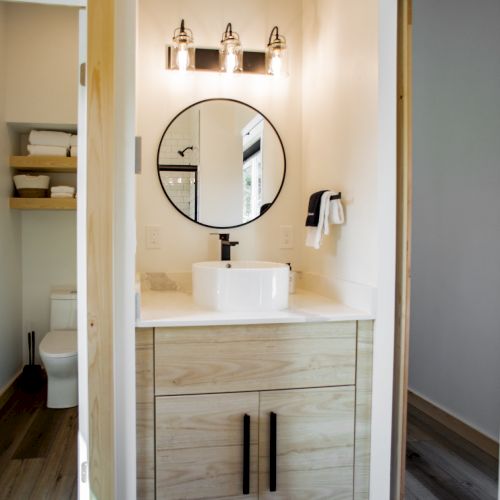 The image shows a bathroom vanity with a round mirror, modern light fixture, and a vessel sink atop a wooden cabinet with dark handles.