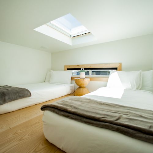 A minimalist bedroom with two beds, soft white linens, brown throws, a small nightstand, and a skylight, creating a serene atmosphere.