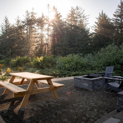 A picnic table and chairs surround a fire pit outdoors, with tall trees and sunlight filtering through in the background.