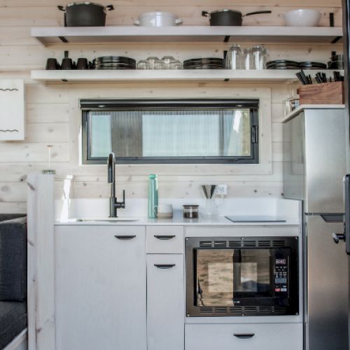 A compact kitchen with white cabinets, a small sink, oven, stovetop, and open shelves holding cookware and dishes, against a wooden wall.