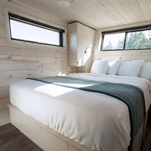 A cozy bedroom with wood-paneled walls, a large bed with white bedding, windows, and a green throw blanket across the foot of the bed.