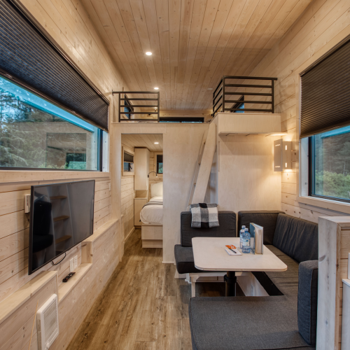 The image shows the interior of a modern tiny house with wood paneling, a loft bed, sofa, TV, and kitchen area. Cozy and efficiently designed.